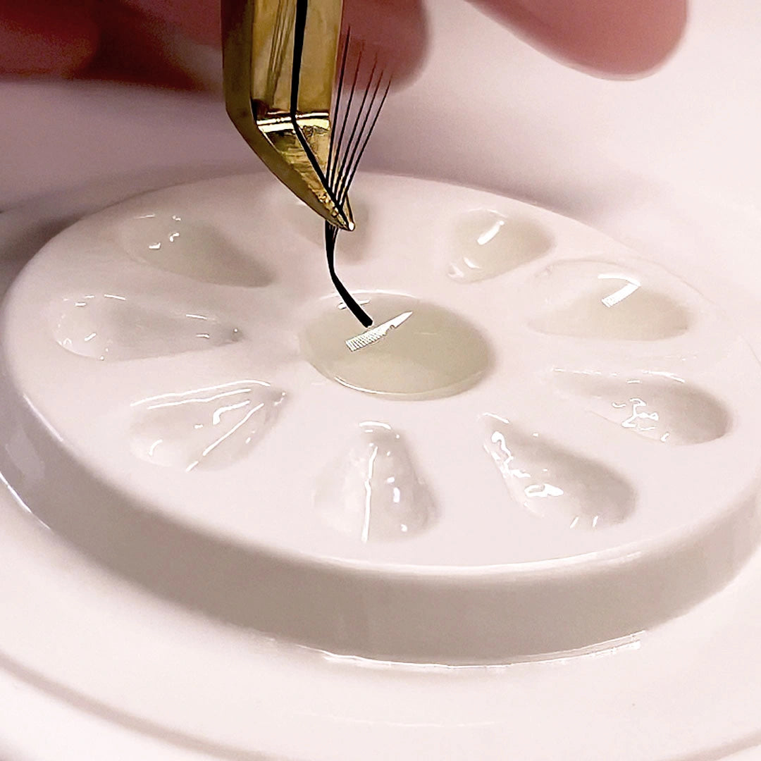 Fotografía macro de unas pinzas doradas sumergiendo la base de un abanico de volumen para extensiones de pestañas en adhesivo transparente sobre una paleta de adhesivo blanca.