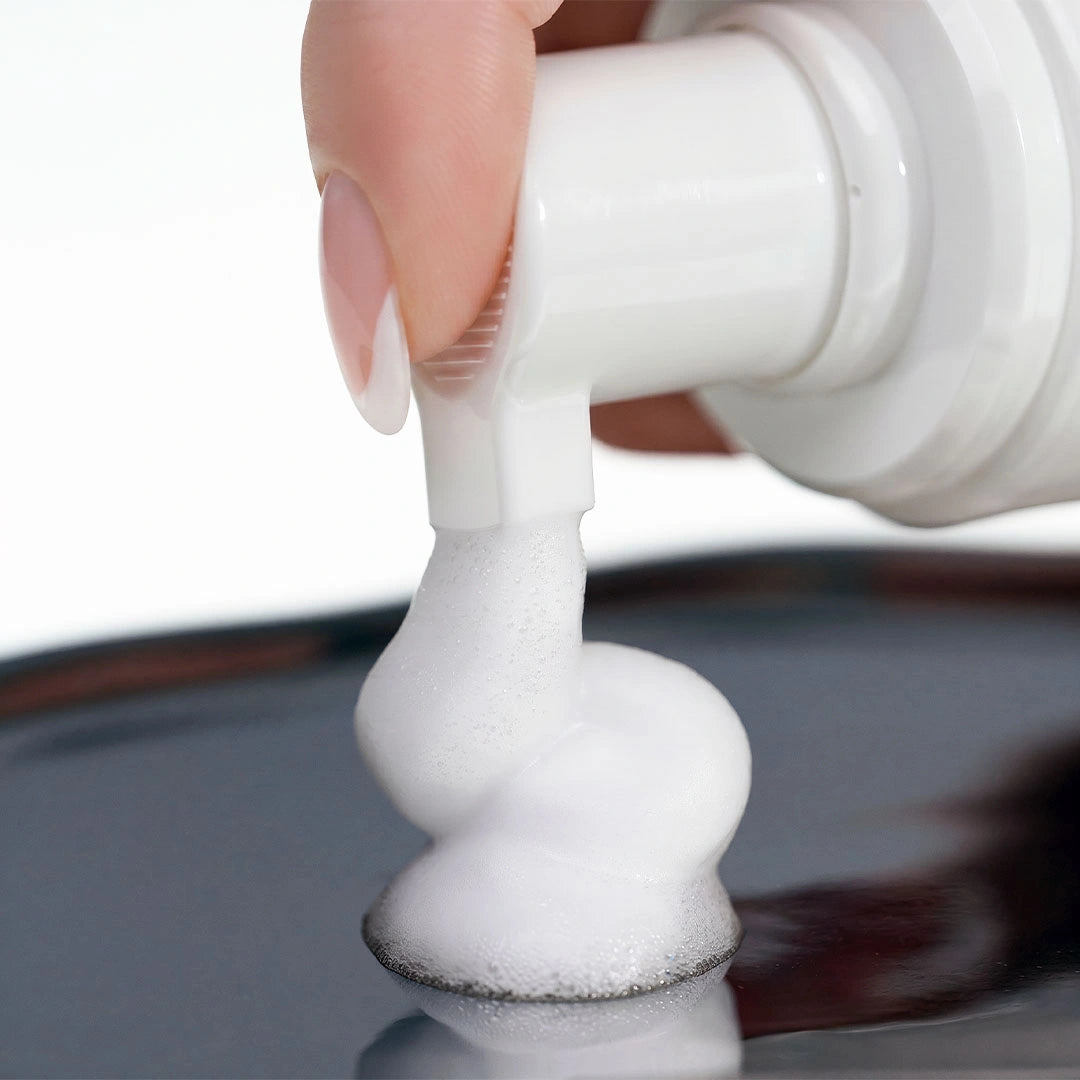Primer plano del dosificador liberando una espuma limpiadora blanca y densa sobre una superficie metálica reflectante.